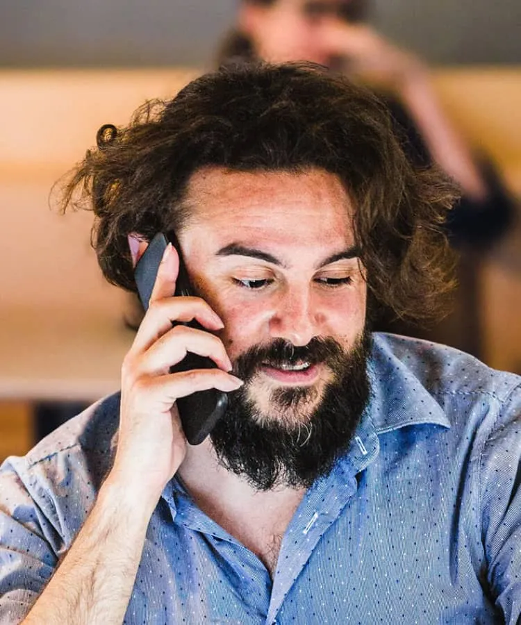 A bearded man sitting at his laptop while talking on the phone