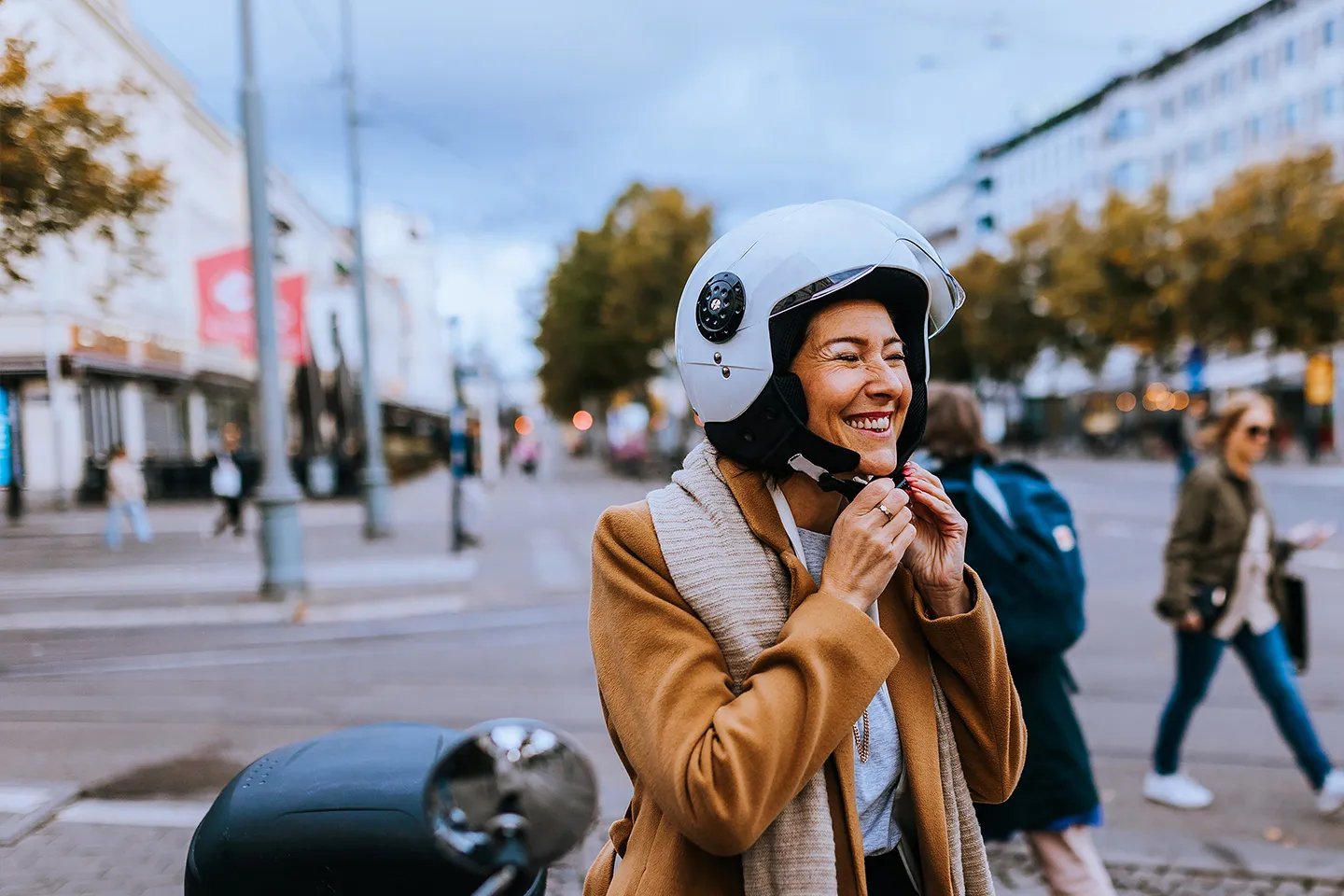 Smiling woman in city center unclipping her motorcycle helmet whilst stood next to her scooter