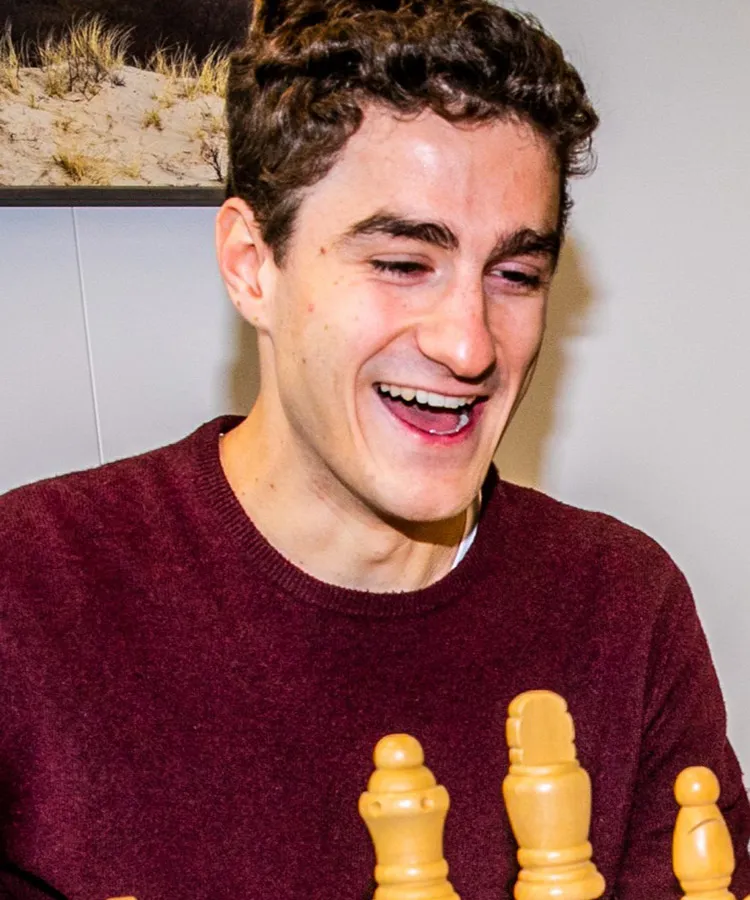 A young man in a burgundy jacket smiling at a chess board
