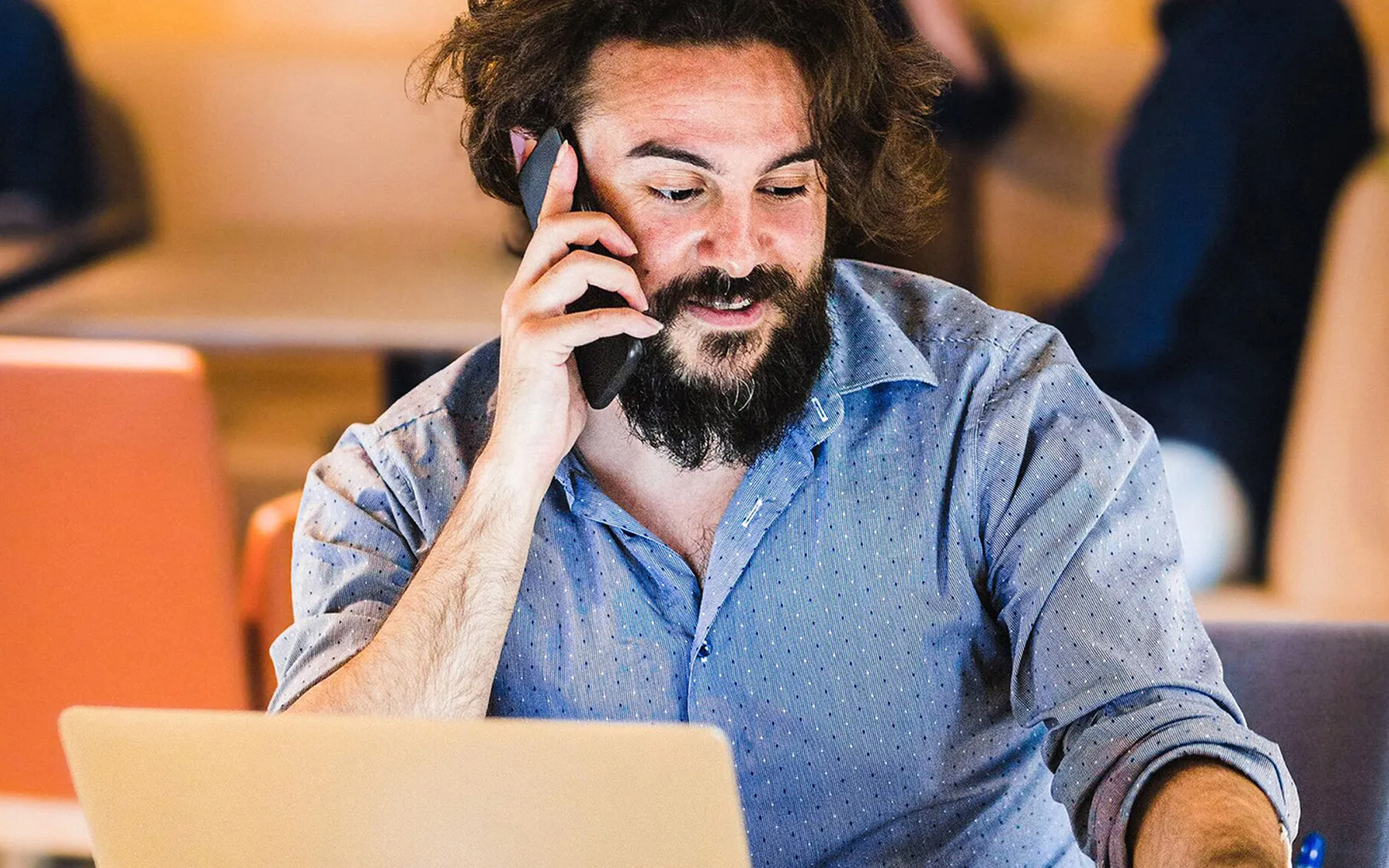 A bearded man sitting at his laptop while talking on the phone
