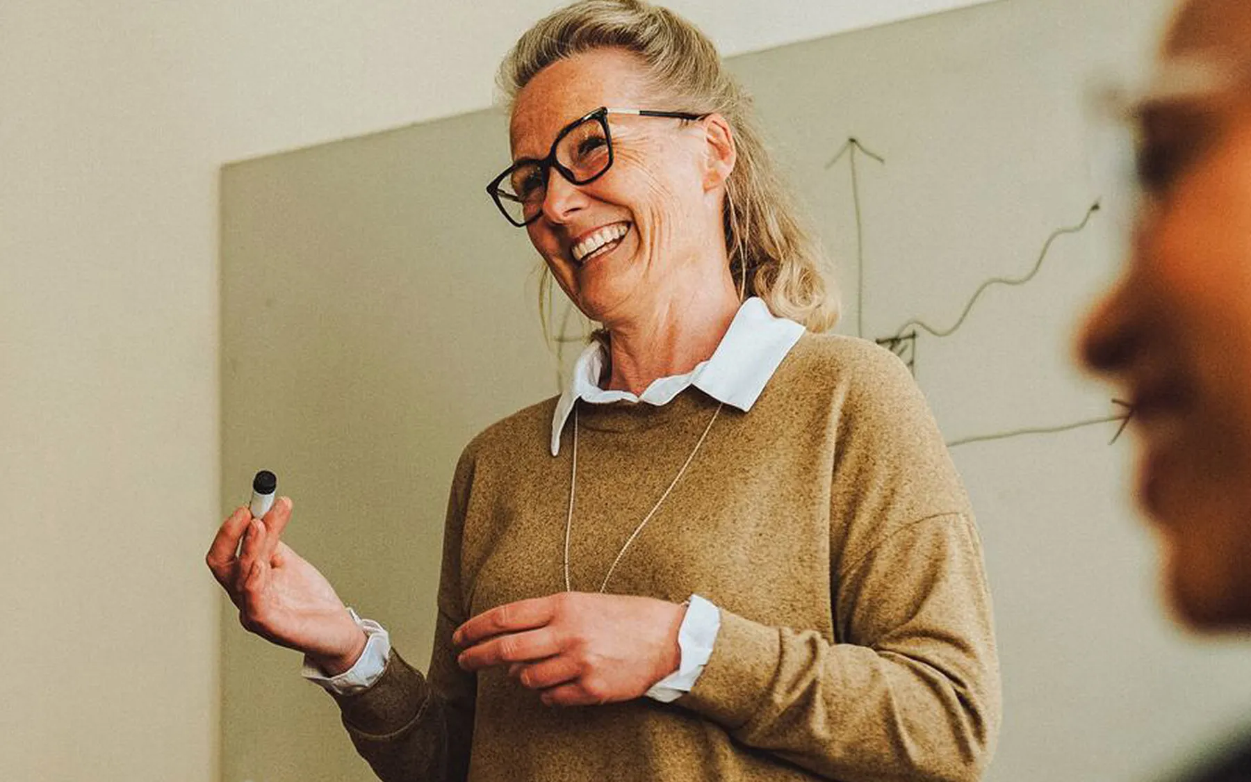 Woman with glasses holding a pen and standing in front of a white board