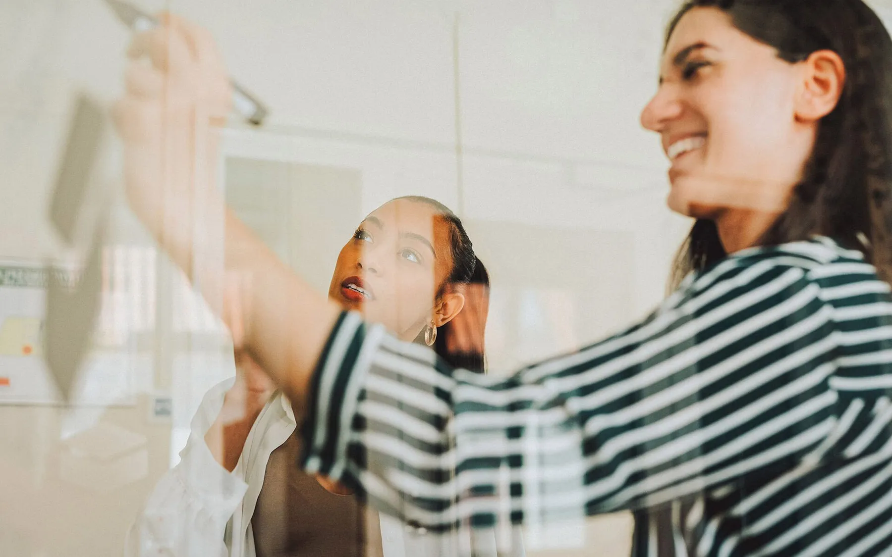 Two young women seen writing on a white board through a glass door
