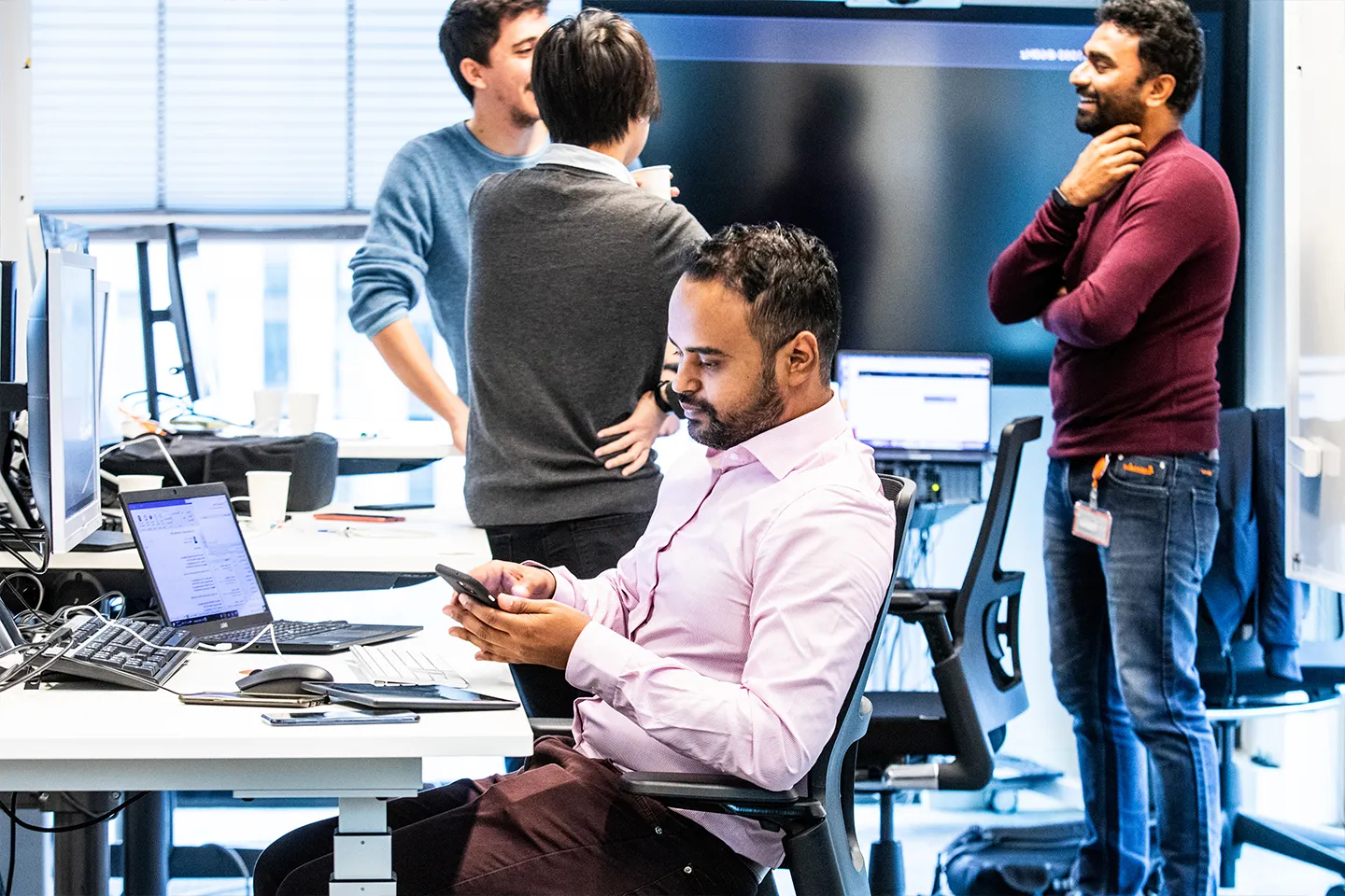 An Asian man sat at his desk, working on his mobile phone with colleagues talking in the background