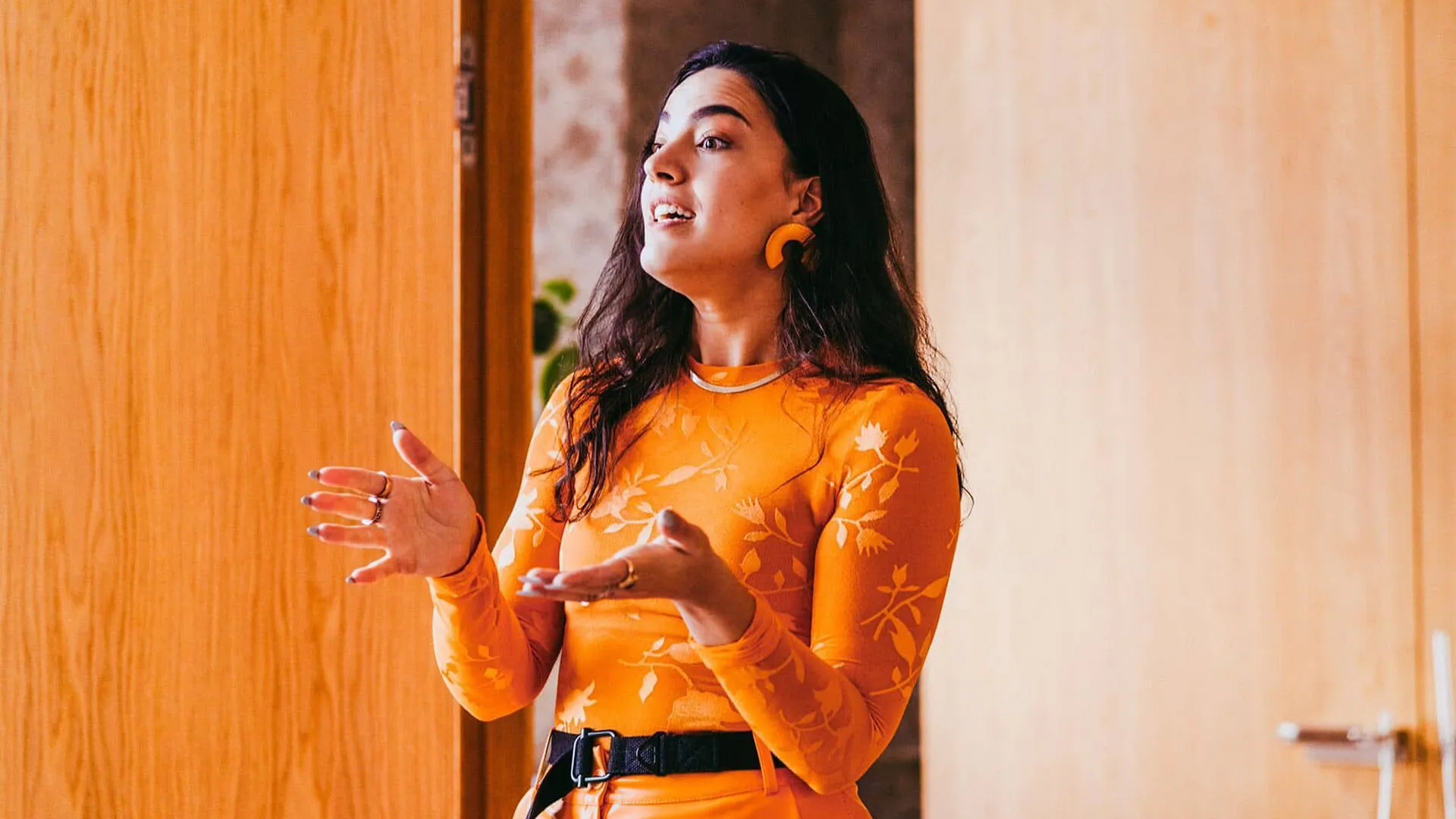 A woman dressed in orange addresses a room with hands outstretched