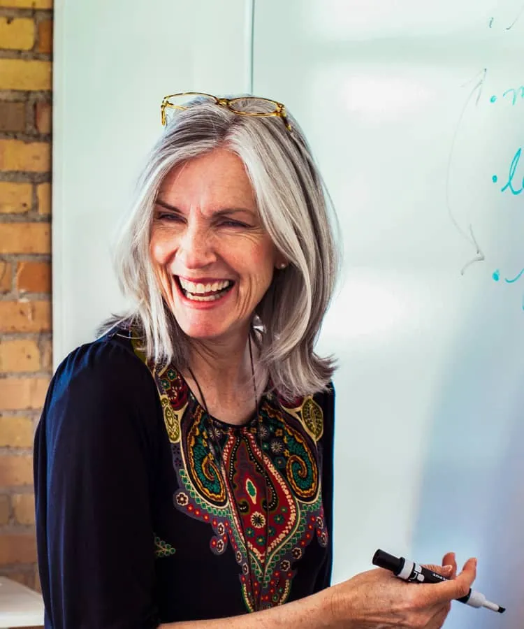 A smiling, grey-haired woman writing on a white board